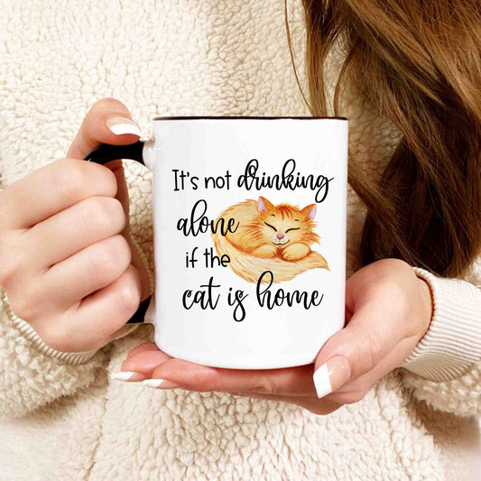 A closeup shot of a woman holding A white coffee mug that reads "It's not drinking alone if the cat is home" in a script font.