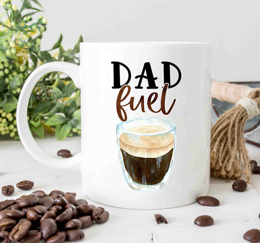 A white coffee mug that reads dad fuel in script font styled on a table with greenery in the background and coffee beans in the foreground.