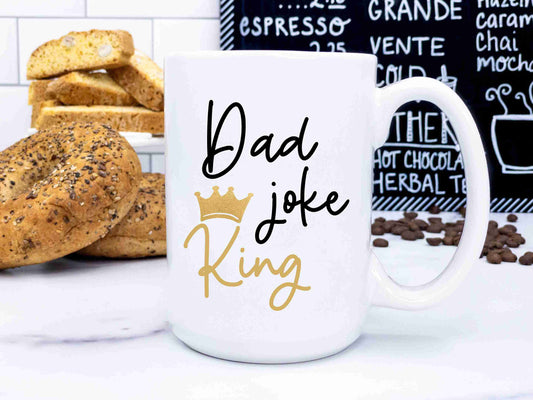 A large coffee mug that reads "dad joke king" in a script font styled in front of baked goods and a coffee shop sign.