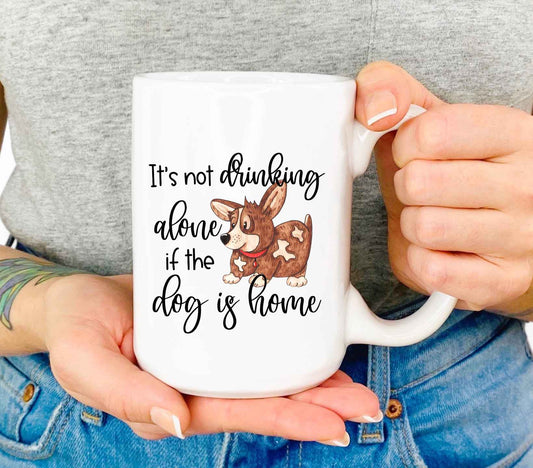 A closeup shot of a woman holding a white coffee mug that reads "it's not drinking alone if the dog is home" in a script font.