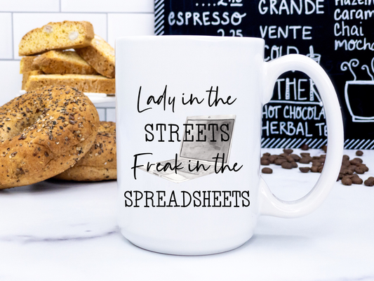 A white coffee mug with a black handle that reads "lady in the streets, freak in the spreadsheets"