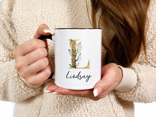 A closeup shot of a woman holding a white coffee mug with a golden L initial with the name Lindsay written underneath.