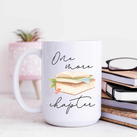 A white coffee mug that reads one more chapter in script font. The mug is staged with a stack of books and a pink plant pot in the background.