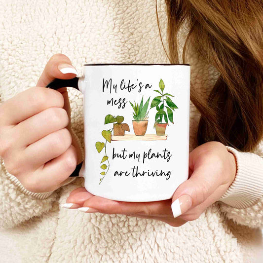 A closeup shot of a woman holding a white coffee mug with a picture of houseplants that reads "My life's a mess but my plants are thriving." in a script font.
