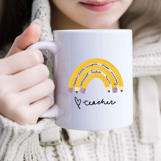 A woman holding a white mug with a rainbow made of pencils interspersed with motivational sayings. The mug has a hand drawn heart and teacher in a script font on the bottom.