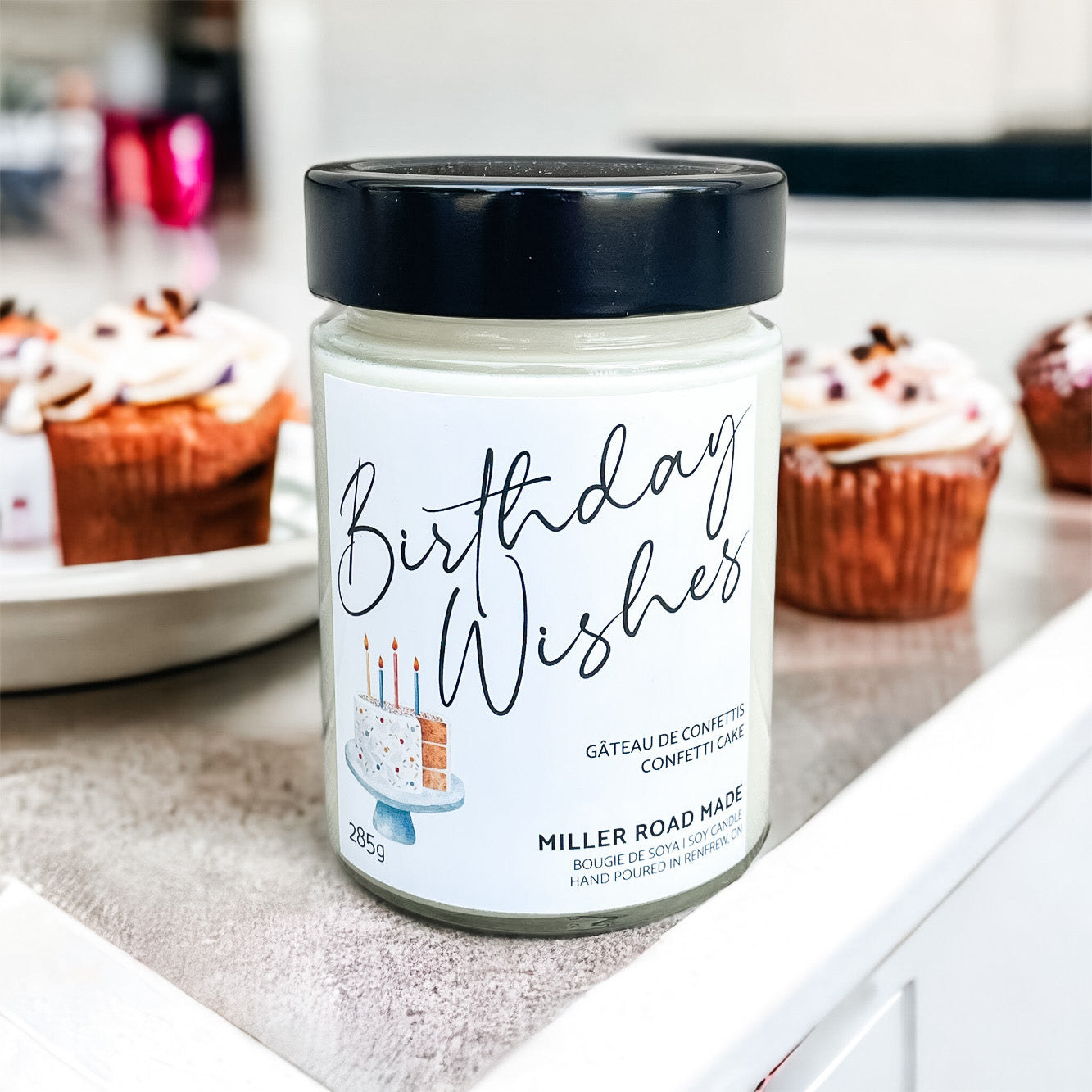 Best candle present. A birthday cake scented candle staged on a counter with cupcakes in the background.