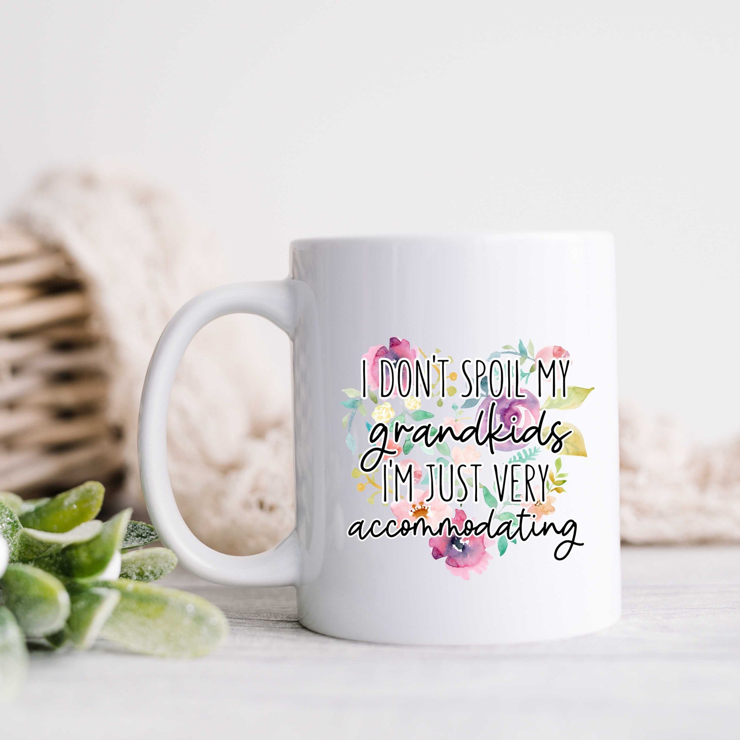 A white mug with a floral print and cursive writing reading I don't spoil my grandkids, I'm just very accommodating.